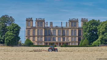 Hardwick Hall tractor This landscape photograph captured in the late morning during the summer features Hardwick Hall, an Elizabethan manor house located in Derbyshire, United Kingdom. The imposing architecture of Hardwick Hall, a well-known National Trust property, dominates the scene with its extensive windows and ornate rooftop details. In the foreground, a red tractor is positioned on the hay-strewn grounds, highlighting rural life in the English countryside. Trees frame the stately manor, reflecting the verdant environment typical of summer. The image showcases the historic and architectural significance of Hardwick Hall, an iconic example of Elizabethan design and one of the most important manor houses in Derbyshire.